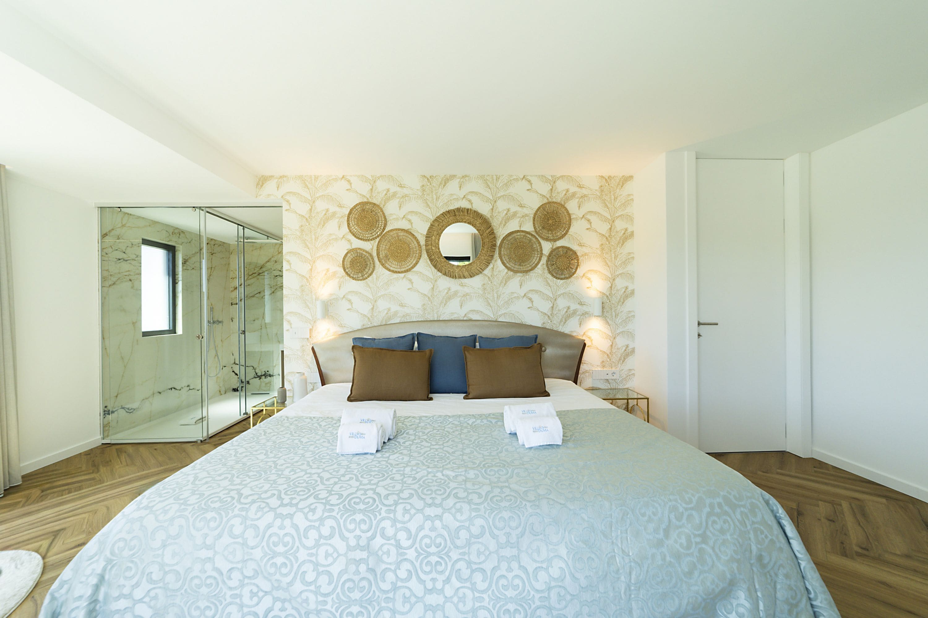 A bedroom with a large bed, a glass shower enclosure, a wall-mounted mirror, and a patterned wall.