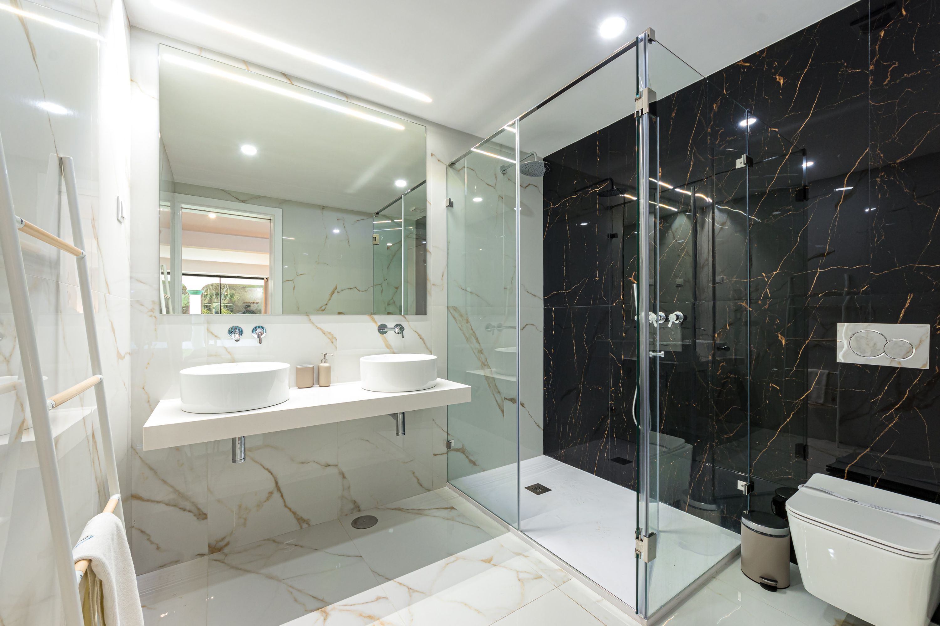 Un baño moderno con ducha de vidrio, dos lavamanos y piso de mármol.
