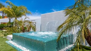 A hot tub with a waterfall feature, surrounded by palm trees and a grassy area. - Casinha in Albufeira (Albufeira)