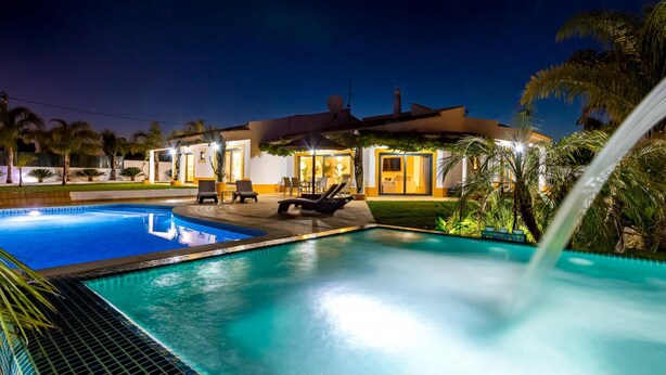 A swimming pool with lounge chairs and a waterfall feature, surrounded by palm trees and a well-lit house. - Casinha in Albufeira (Albufeira)