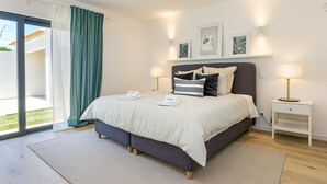 A modern bedroom with a large bed, bedside lamps, a small bedside table, and a view through a glass door. - Casinha in Albufeira (Albufeira)
