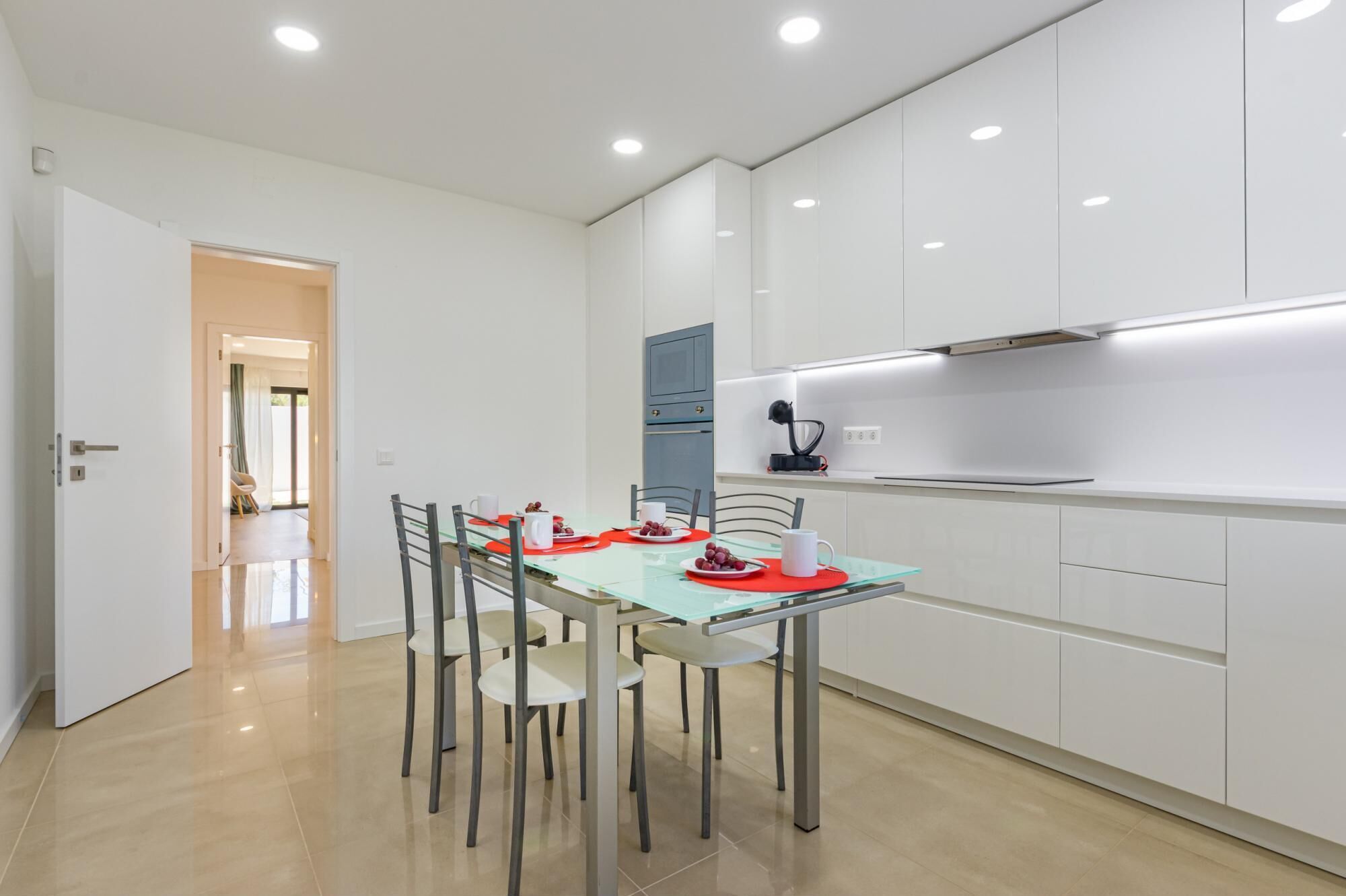 Una cocina moderna con armarios blancos, una mesa de comedor de vidrio y un mantel individual rojo.