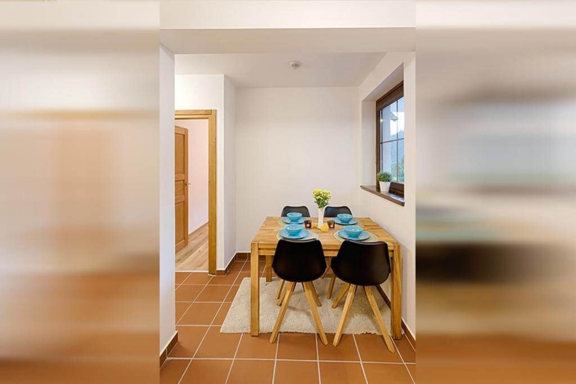 A dining area with a wooden table, four black chairs, and a small potted plant on the windowsill.