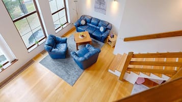 Amplia sala de estar con un sofá de cuero azul, una mesa de centro de madera y un tapete gris. Se observa una escalera de madera al frente.