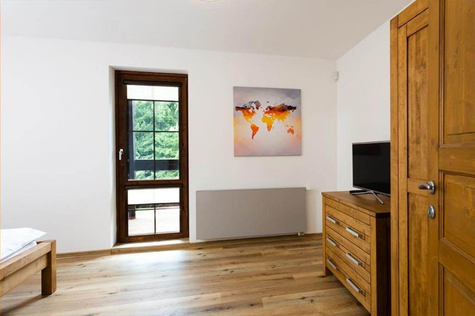 A room with a wooden floor, a wooden cabinet with drawers, a television on top, a window with a view of greenery, and a world map painting on the wall.