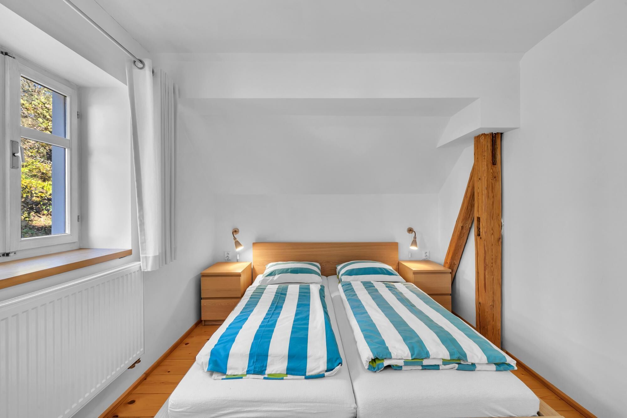 A small, bright room with two single beds, a window, and a radiator.