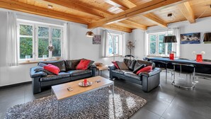 Un salon moderne avec deux canapés en cuir, une table basse en bois et un coin repas comprenant une table et des chaises. De grandes fenêtres laissent entrer la lumière naturelle et offrent une vue sur la verdure.