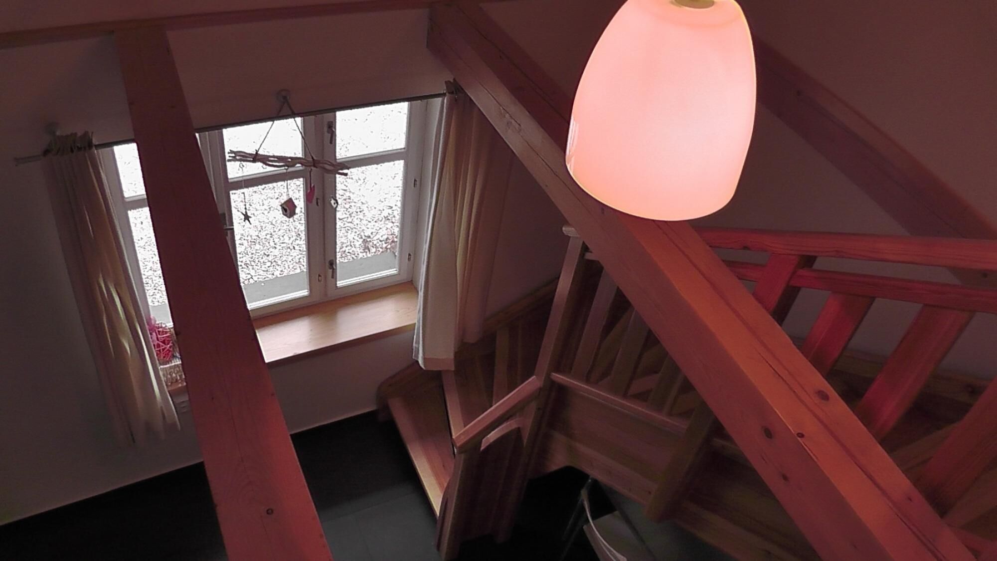 A room with a wooden staircase, a window with curtains, and a hanging light fixture.
