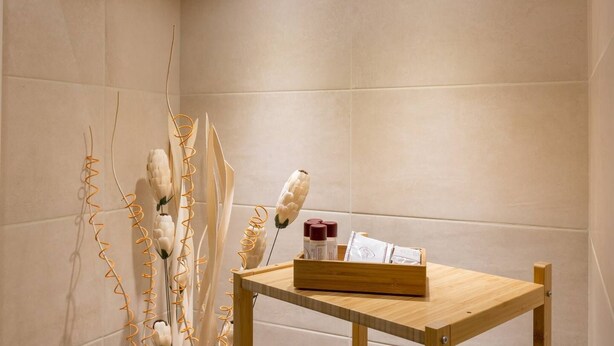 Une salle de bain carrelée de carreaux beige, une table en bois sur laquelle est posée une corbeille contenant des articles de toilette, et des fleurs blanches décoratives aux tiges orange.