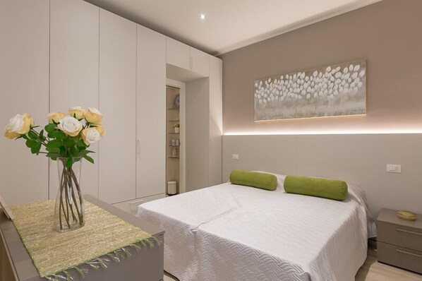 Une chambre à coucher comprenant un lit, une table de chevet, une commode, un vase avec des fleurs et une œuvre d’art accrochée au mur.