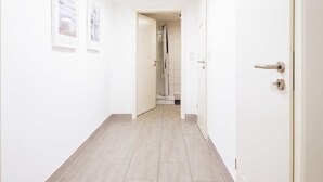Un pasillo limpio y moderno con paredes blancas y suelo de madera clara. Se observan cuadros enmarcados en la pared, una puerta a la derecha y una zona de ducha al fondo.