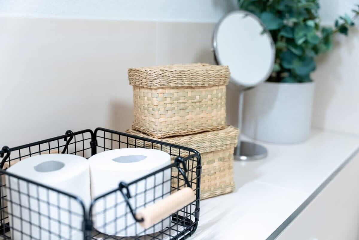 Een badkamer met een witte wastafel, een zwarte metalen mand met toiletpapier en twee gevlochten manden.