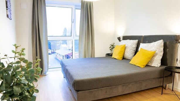 Een moderne slaapkamer met een groot bed, grijze beddengoed en gele kussens. Links staat een potplant en er is een raam met gordijnen.