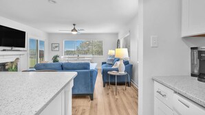 A modern living room with a flat-screen TV, blue sofas, a small round table with a lamp, and a kitchen area with a coffee maker.