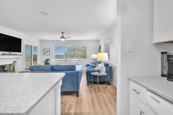 A modern living room with a flat-screen TV, blue sofas, a small round table with a lamp, and a kitchen area with a coffee maker.