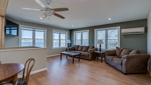 A spacious living room with a ceiling fan, large windows, a brown leather couch, a wooden coffee table, and a dining area with wooden chairs.