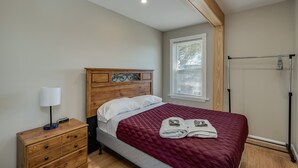 A bedroom with a wooden headboard, a bed with a red quilt, a nightstand with a lamp, and a window with blinds.