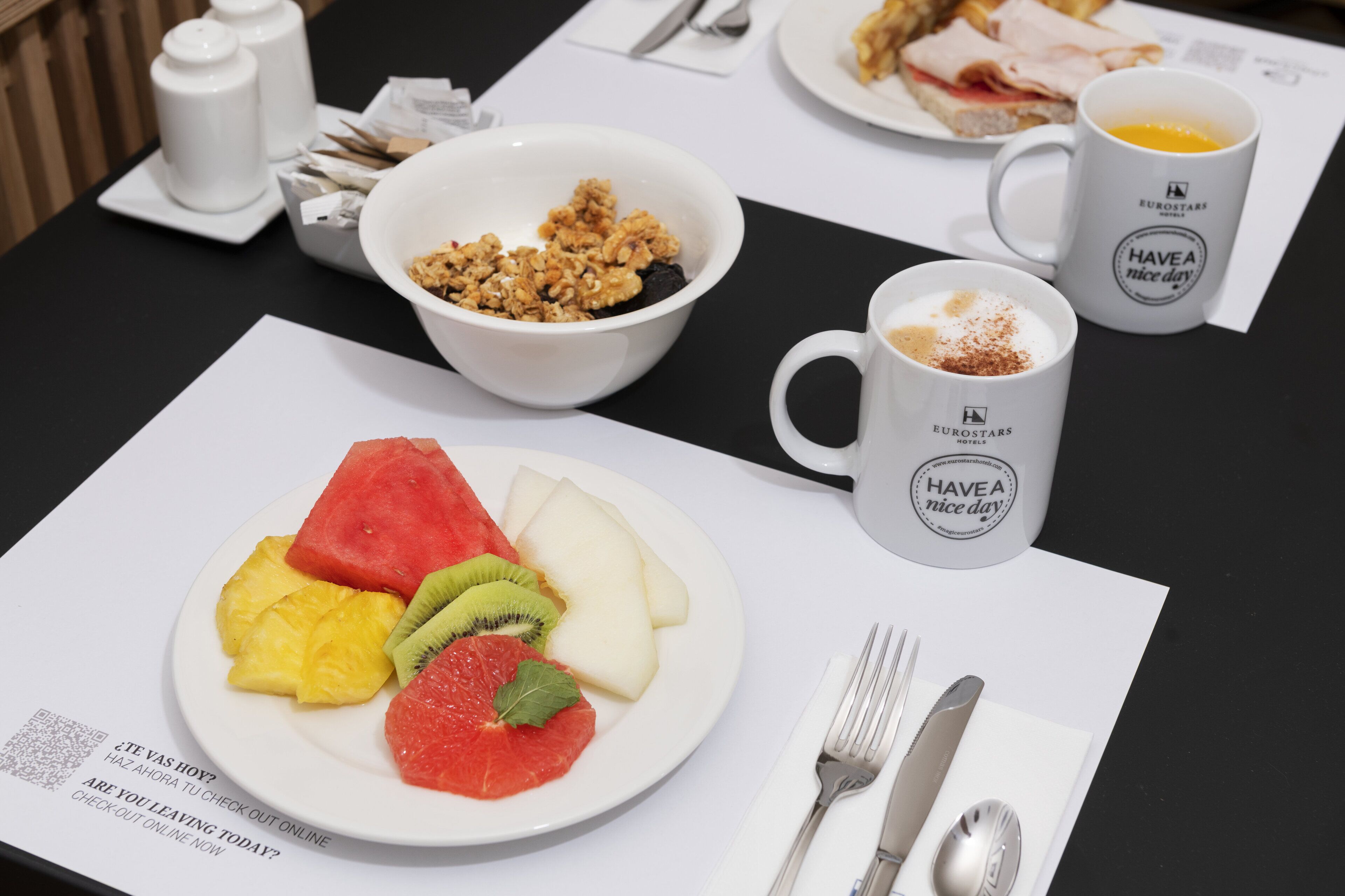 Frühstückstisch mit Obstteller, einer Schüssel Granola und zwei Tassen mit der Aufschrift 'HAVE A nice day'.