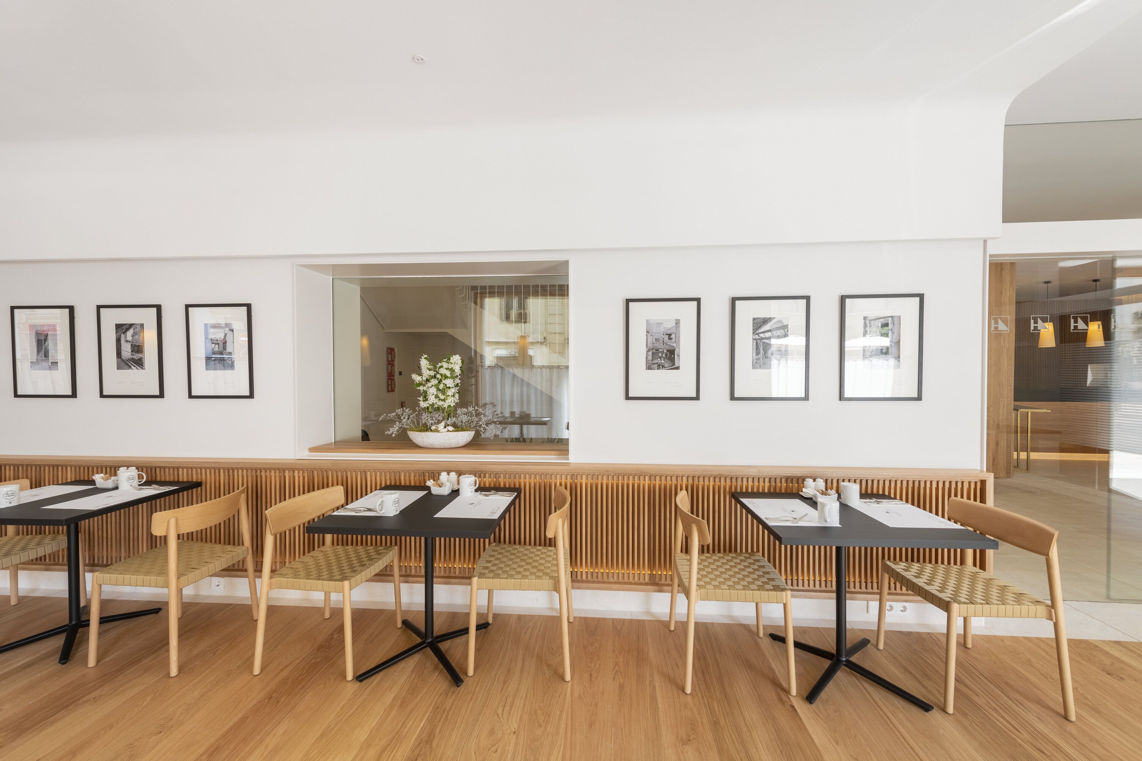 Un interior moderno con suelo de madera, mesas y sillas de madera y color negro. Hay cuadros enmarcados en las paredes y una ventana con vistas al exterior.