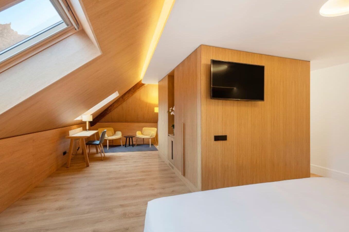 Een moderne, compacte kamer met een schuine kap, een flatscreen televisie aan de wand, een klein zithoekje met een tafel en stoelen, en een bed met een wit hoofdeinde.
