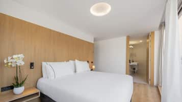 A modern hotel room with a large bed, wooden headboard, bedside table with a plant, and a bathroom visible through an open door.