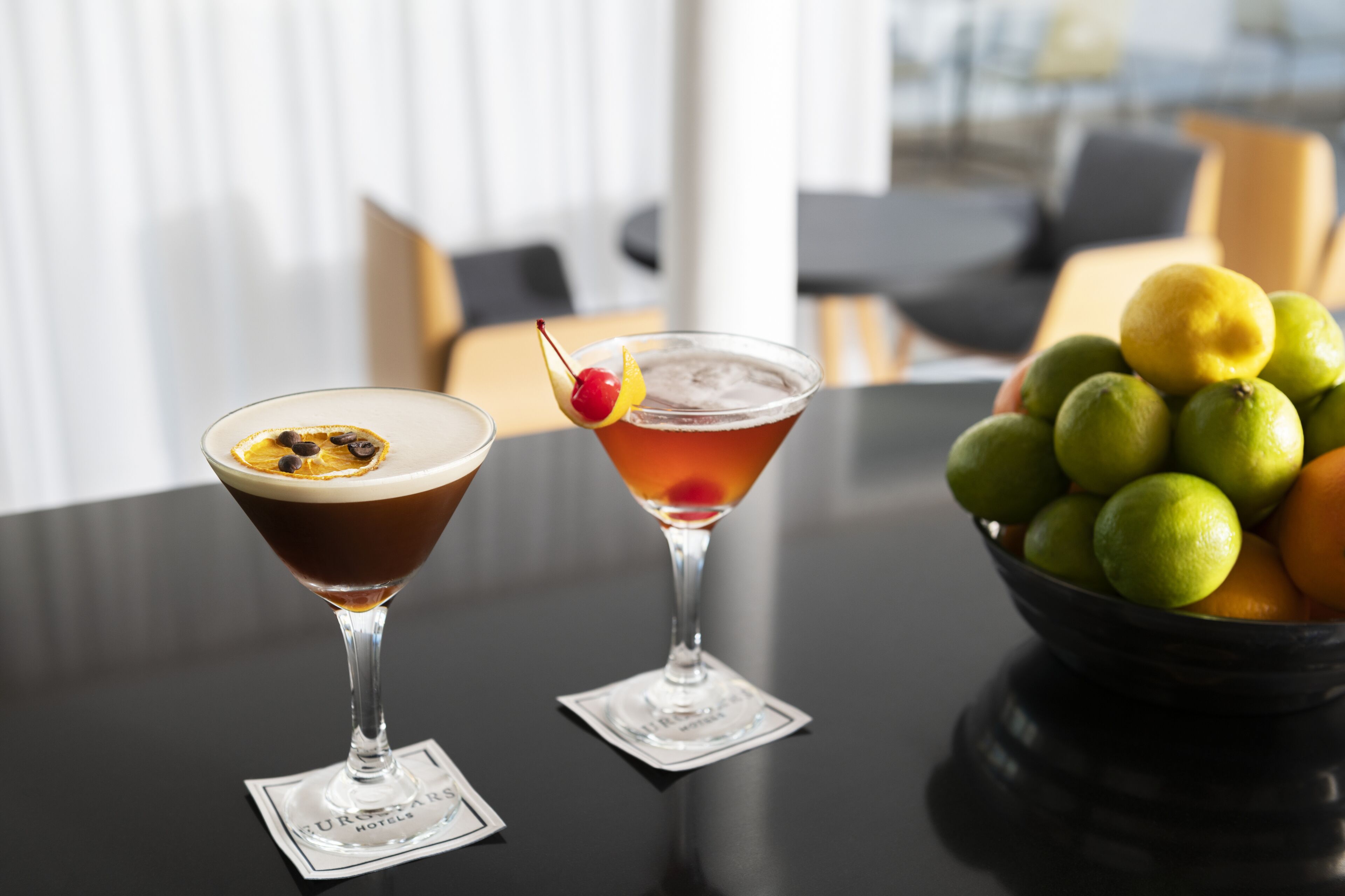 Dos vasos de martini sobre una mesa, junto a un cuenco de limones y naranjas.