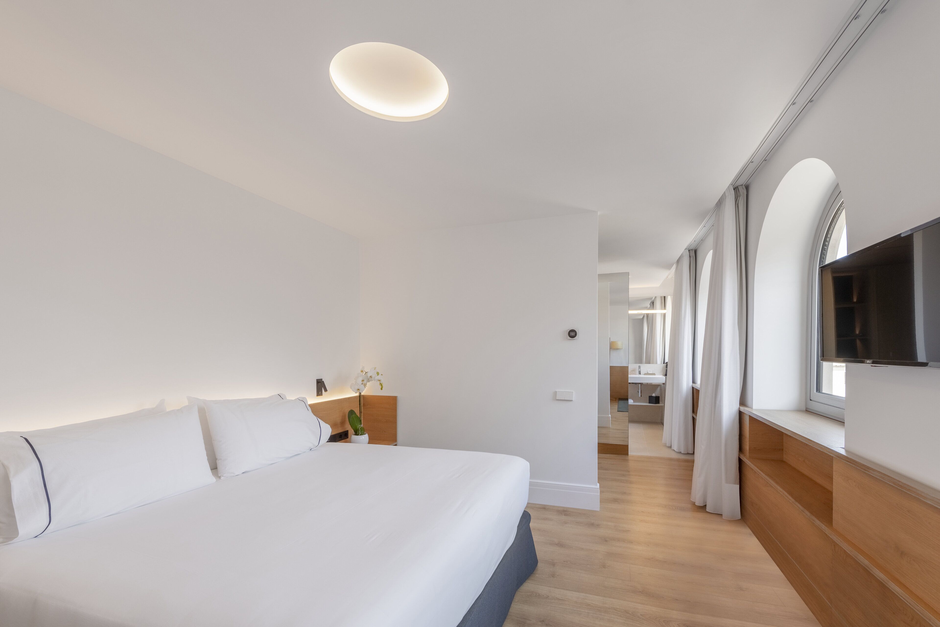 Una habitación de hotel moderna con una cama grande, un televisor de pantalla plana montado en la pared y un banco de madera junto a la ventana.
