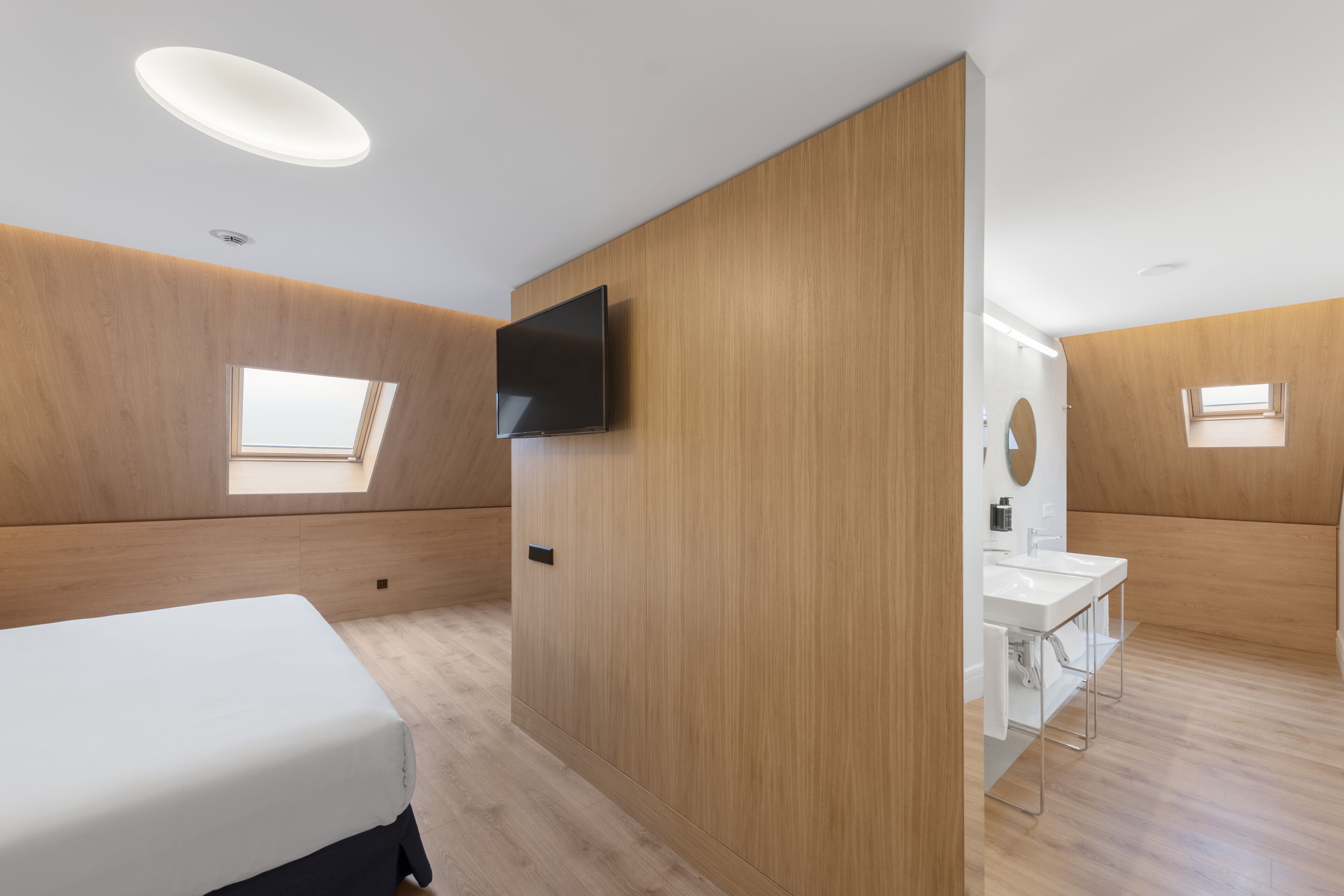 A modern bedroom with a bed, a TV, a skylight, and a bathroom with a sink and mirror.