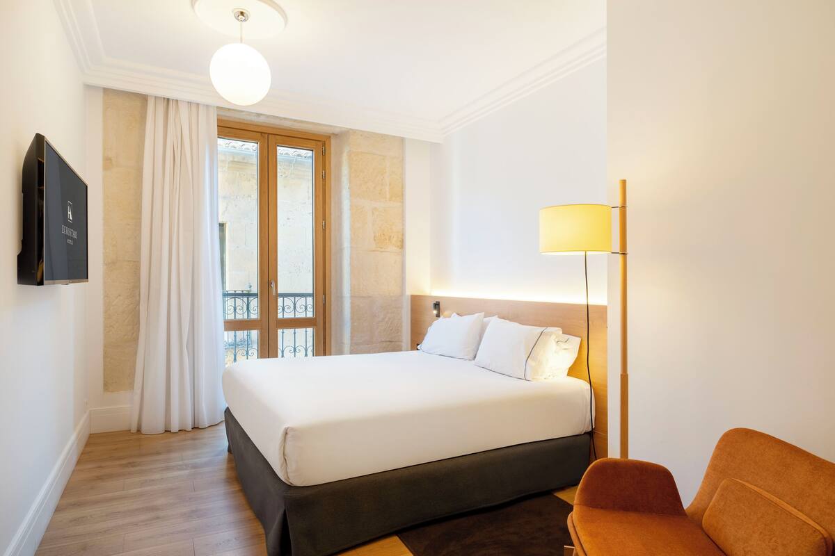 A hotel room with a bed, a chair, a TV, a window with curtains, and a lamp.