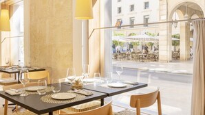 Un restaurante con mesas preparadas para comer, vista a la calle y otros establecimientos de comida, y un interior con pared de piedra.