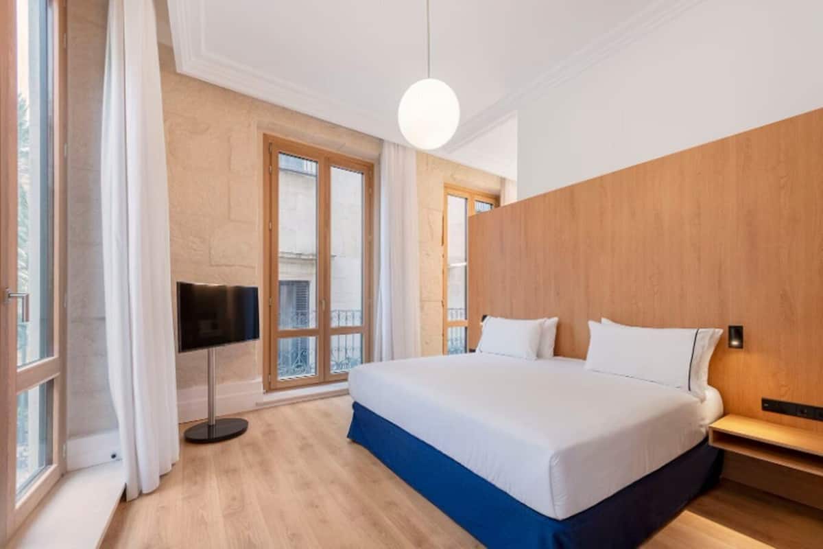 A modern hotel room with a large bed, wooden paneling, a TV, and a balcony view.