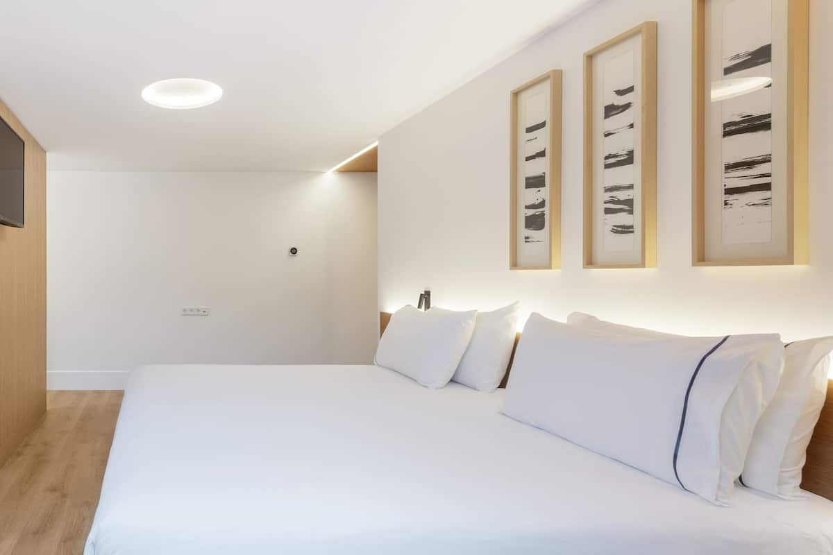 A modern hotel room with a large bed, wooden paneling, a flat-screen TV, and framed artwork on the wall.