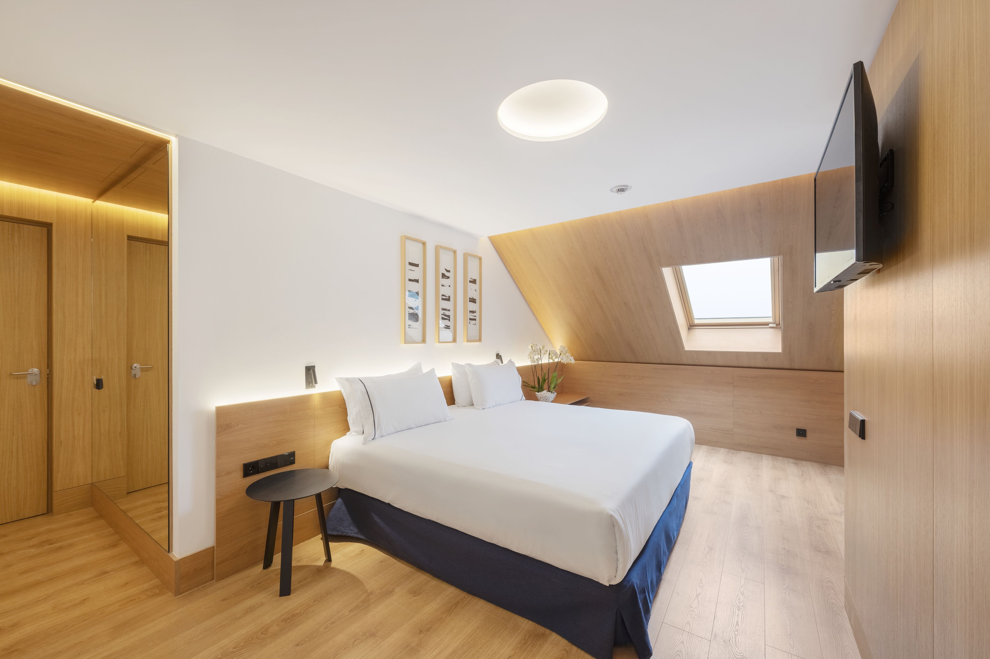 A modern bedroom with a large bed, a small table, a TV mounted on the wall, and a skylight.