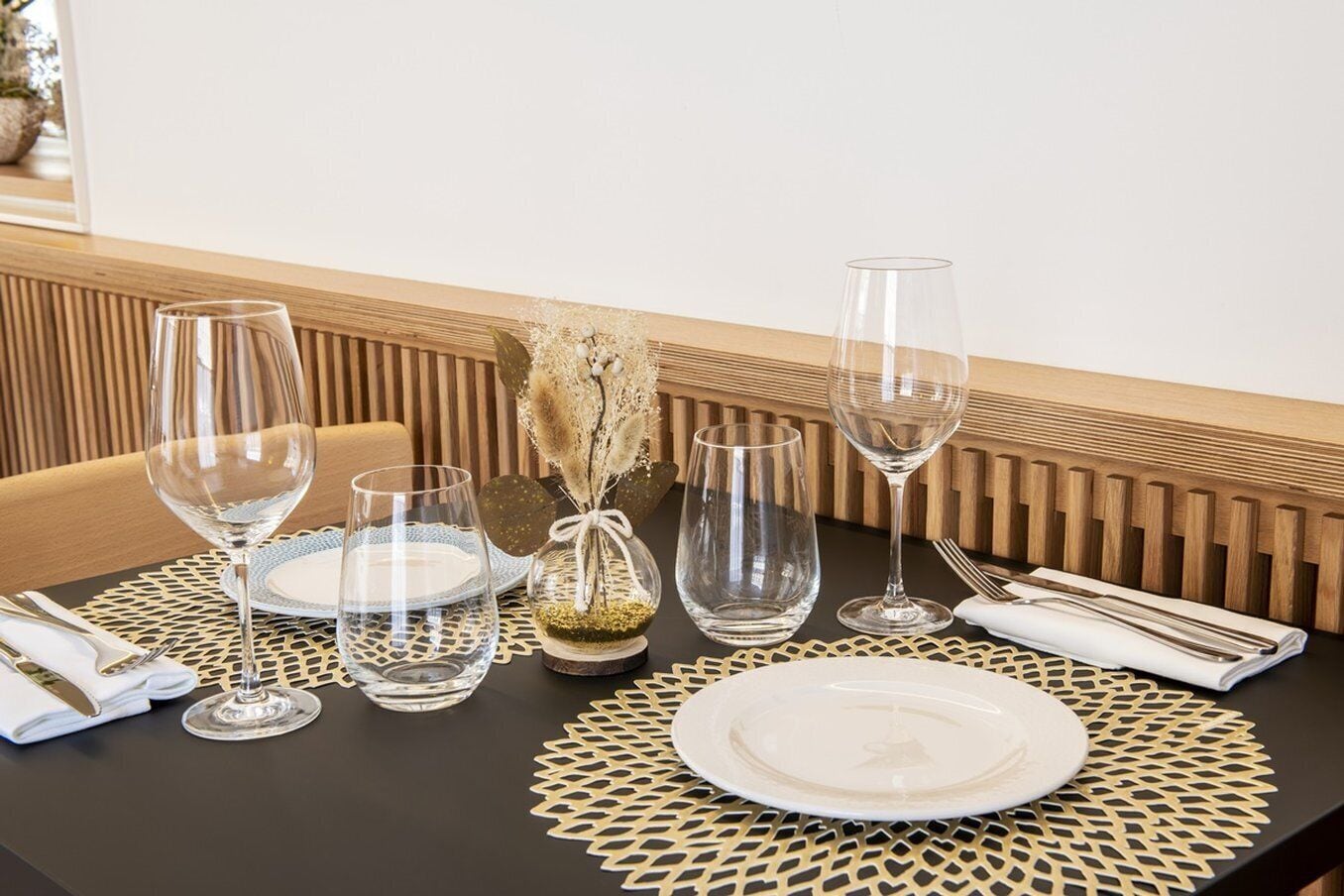 Mesa de jantar posta com taças de vinho, pratos e talheres, apresentando um centro de mesa decorativo e um sousplat texturizado.