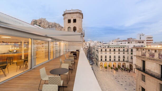 Una terraza en la azotea con mobiliario exterior, vistas a la ciudad y una torre histórica al fondo.