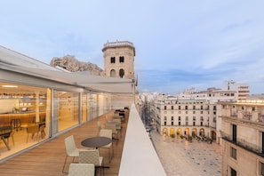 Una terraza en la azotea con mobiliario exterior, vistas a la ciudad y una torre histórica al fondo.