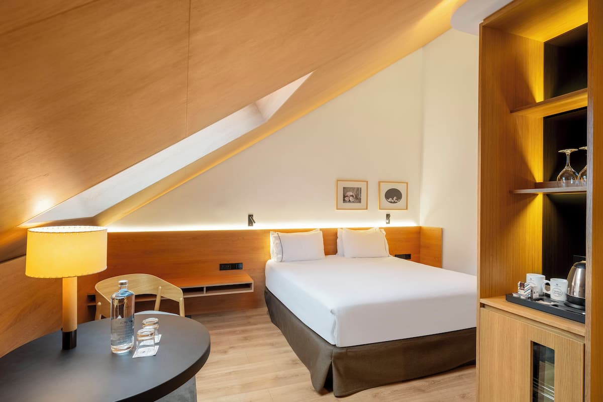 A modern hotel room with a bed, a desk, a lamp, and a shelf with glasses and a kettle.