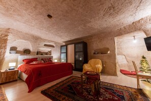 A bedroom with a red bed, a yellow armchair, a small table, a Christmas tree, and a stone wall. - Cappadocia Deep Cave House (Ürgüp)