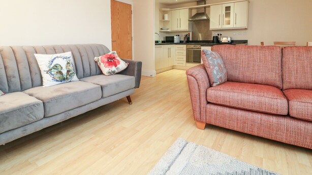 A living room with two sofas, a kitchen in the background, and a wooden floor. - 1 Tregonwell Court (Minehead)