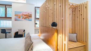 A modern bedroom with a wooden headboard, a bed with white linens, a striped pillow, a desk with a chair, and a painting of a bridge. - Design Rooms Verona (Verona)