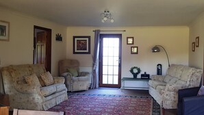 Sala de estar com dois sofás de padrão floral, um banquinho, um abajur e uma porta que dá para outro quarto.