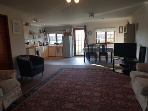 A spacious living area with a large rug, a dining table with chairs, a television, and a kitchen in the background. - Spacious pet-friendly, accessible family holiday home at Hawthorn Farm, Kent (Ware, Ash, Canterbury)