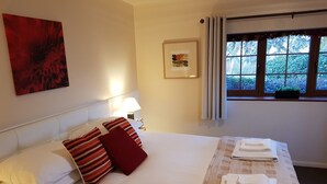 Quarto de hotel com uma cama grande, uma mesa de cabeceira com abajur, uma janela com cortinas e um quadro na parede.