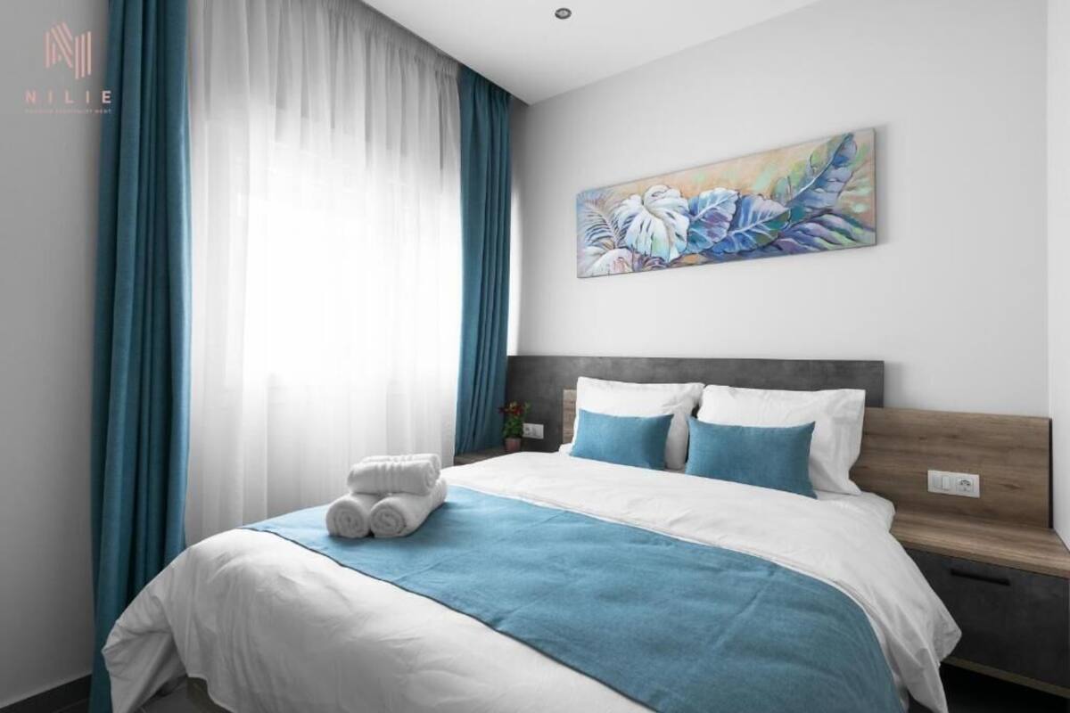 Une chambre d’hôtel avec un lit, une literie bleue et blanche, un tableau au mur et une fenêtre avec des rideaux.