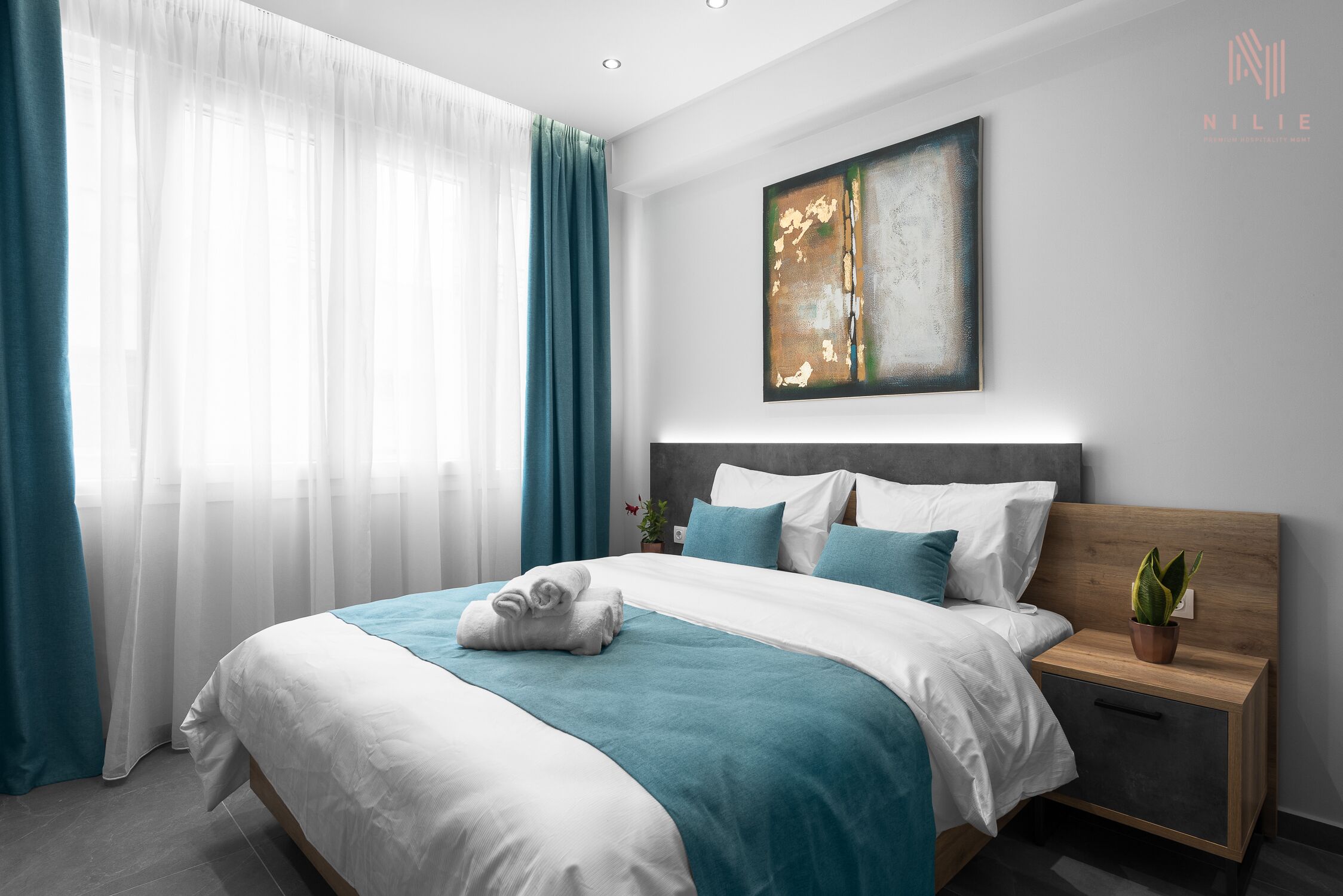 A modern hotel room with a neatly made bed, a bedside table with a plant, and a large abstract painting on the wall.