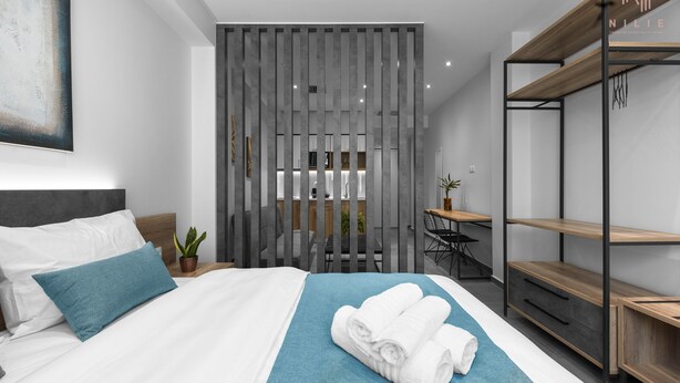 A modern hotel room with a neatly made bed, a blue accent pillow, a set of towels, a desk with a chair, and a wooden shelving unit. - Factory Inn Nilie Hospitality MGMT (Thessaloniki)
