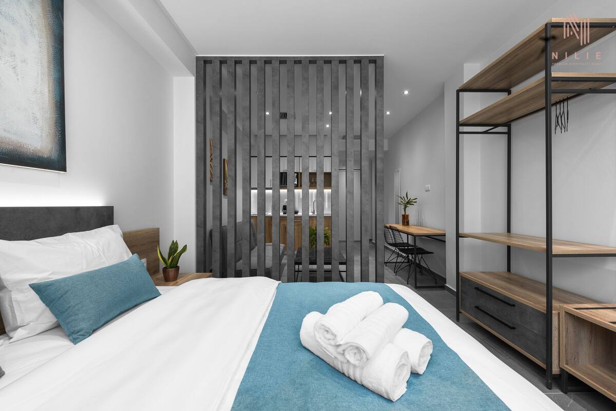 Une chambre d’hôtel moderne avec un lit soigneusement fait, un coussin bleu décoratif, un ensemble de serviettes, un bureau avec une chaise et une étagère en bois.