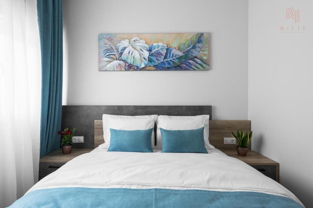 Un dormitorio con cama, dos mesitas de noche, un cuadro en la pared y una ventana con cortinas.