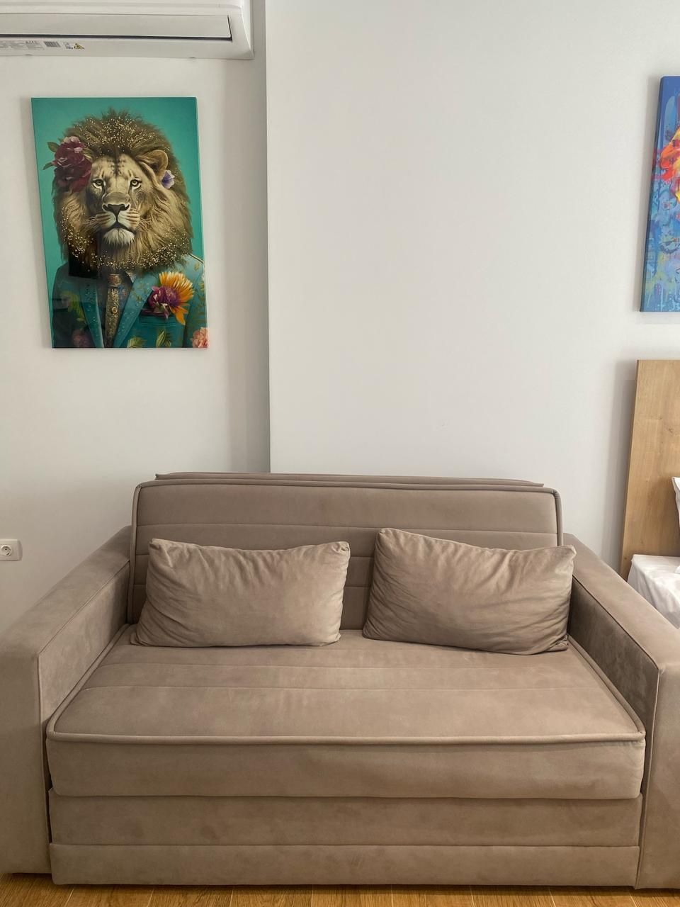 A beige sofa with two cushions, a teal painting of a lion, and a wooden headboard.