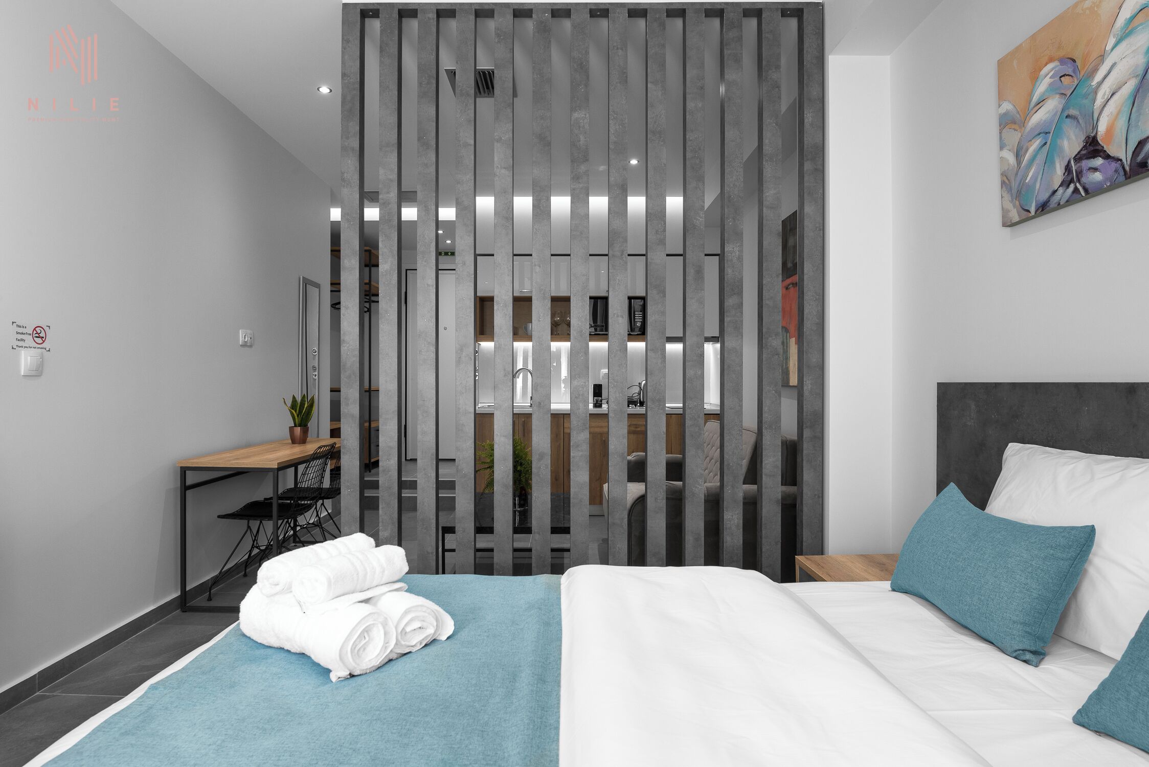 A modern hotel room with a bed, a desk, a chair, and a bathroom visible through a partition.